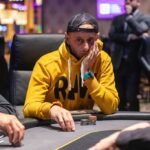 Ein Mann und eine Frau spielen Poker an einem eleganten Holztisch in einem stilvollen Raum mit Kronleuchter. Die Frau hat ein Glas Wein in der Hand, während der Mann konzentriert seine Karten betrachtet.