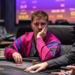 Ein Mann und eine Frau spielen Poker an einem Tisch in einem eleganten Casino. Der Mann hat Karten in der Hand und der Blick der Frau ist konzentriert. Im Hintergrund sind weitere Spieler und eine glamouröse Einricht