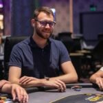 Ein Gruppenfoto von vier Freunden an einem Pokertisch, die Karten halten und lächeln. Ein Mann mit dunkler Brille und Kappe mischt Karten in der Mitte des Tisches. Hinter ihnen sind Poker