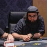 Ein Mann und eine Frau sitzen an einem Pokertisch und spielen Karten. Der Mann hat eine Hand voller Chips, während die Frau konzentriert ihre Karten betrachtet. Die Szene spielt sich in einem eleganten