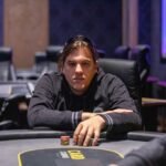 Ein Mann in einem schwarzen Anzug und mit einer Sonnenbrille sitzt an einem Pokertisch und betrachtet seine Karten. Andere Spieler und Dealer sind im Hintergrund zu sehen.
