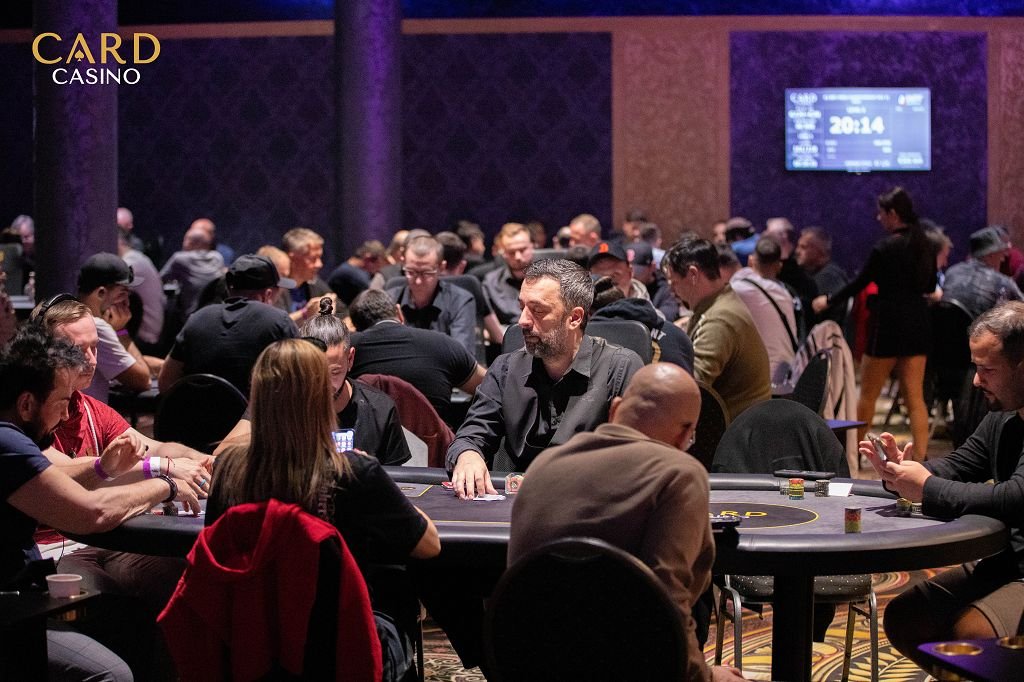 Ein Mann und eine Frau sitzen an einem Pokertisch und spielen ein Kartenspiel. Der Mann hat einen ernsten Gesichtsausdruck und hält Karten in der Hand, während die Frau ihn konzentriert ansie