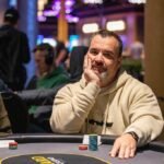 Ein Mann in einem schwarzen Anzug und einer Sonnenbrille sitzt an einem Pokertisch und hält zwei Spielkarten in der Hand. In der Mitte des Tisches liegen weitere Karten und Pokerchips.