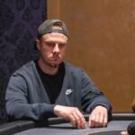 Ein Mann spielt Poker an einem Tisch in einem eleganten Casino, umgeben von anderen Spielern. Er hat eine ernste Miene und hält Karten in der Hand, während er auf seinen Gegner schaut. Die Atmosph