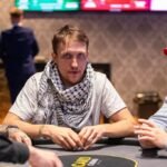 Ein Mann sitzt an einem Pokertisch und hält zwei Karten in der Hand, während er konzentriert auf die Gemeinschaftskarten in der Mitte des Tisches schaut. Andere Spieler sind im Hintergrund