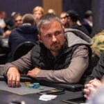 Ein Mann spielt Poker an einem Tisch mit grünem Filz und hält eine Reihe von Spielkarten in der Hand. Im Hintergrund sind weitere Spieler und Chips zu sehen.
