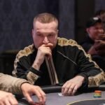 Ein Mann sitzt an einem Pokertisch und hält zwei Spielkarten in der Hand. Er schaut konzentriert auf seine Karten, während im Hintergrund andere Spieler und Chips zu sehen sind.