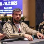 Ein Mann sitzt an einem Pokertisch und hält zwei Karten in der Hand, während er konzentriert auf die Gemeinschaftskarten schaut. Andere Spieler sind im Hintergrund zu sehen.