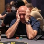 Ein Mann und eine Frau sitzen an einem Pokertisch und spielen Karten. Der Mann hat eine Hand mit Chips und Karten vor sich, während die Frau konzentriert ihre Karten betrachtet. Im Hintergrund sind weitere