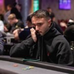 Ein Mann spielt Poker an einem Tisch mit Chips und Karten vor sich. Er hat eine ernste Miene und konzentriert sich auf sein Blatt. Andere Spieler sind im Hintergrund zu sehen.
