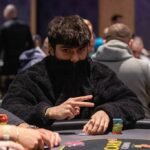Ein professioneller Pokertisch mit grünem Filz, umgeben von Stühlen und Chips. Zwei Spieler mit Karten in der Hand sitzen gegenüber voneinander. Eine spannende Pokerrunde ist im G