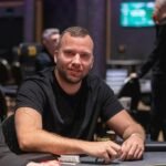 Ein Mann in einem dunklen Anzug und einer Krawatte sitzt an einem Pokertisch und hält zwei Spielkarten in der Hand, während er konzentriert in die Kamera schaut. Im Hintergrund sind weitere