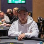 Ein Mann in einem schwarzen Anzug und einer schwarzen Sonnenbrille spielt Poker an einem eleganten Holztisch in einem luxuriösen Casino.