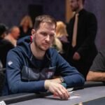 Ein Mann und eine Frau sitzen an einem Pokertisch und spielen Poker. Der Mann hat einen Stapel Chips vor sich, während die Frau ihre Karten betrachtet. Die Szene spielt sich in einem eleganten Raum mit gedimm