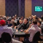 Ein Mann sitzt an einem Pokertisch und hält zwei Spielkarten in der Hand. Er schaut konzentriert auf den Tisch, umgeben von Pokerchips und anderen Spielern im Hintergrund.