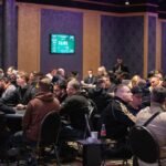 Ein Mann und eine Frau spielen Poker an einem Holztisch in einem gemütlichen Raum mit Teppichboden und warmem Licht. Der Mann hat eine Hand mit Spielkarten geöffnet, während die Frau konzentriert ihre