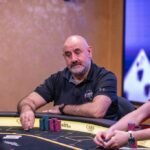 Ein Mann sitzt an einem Pokertisch und hält zwei Spielkarten in der Hand, während er konzentriert auf den Tisch schaut. Um ihn herum sind Pokerchips und Spielkarten verstreut.
