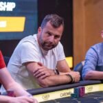 Ein Mann und eine Frau sitzen an einem Pokertisch und betrachten konzentriert ihre Karten. Im Hintergrund sind Pokerchips und Getränke zu sehen.