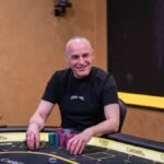 Ein Mann und eine Frau sitzen an einem Pokertisch und spielen Karten. Der Mann hat einen Stapel Chips vor sich und die Frau hält ihre Karten in der Hand. Im Hintergrund sind andere Spieler und eine beleuchtete