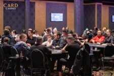Ein Mann in einem schwarzen Anzug sitzt an einem Pokertisch, hält Karten in der Hand und schaut konzentriert auf seine Gegner. Die anderen Spieler sind teilweise im Bild zu sehen, während im Hinter