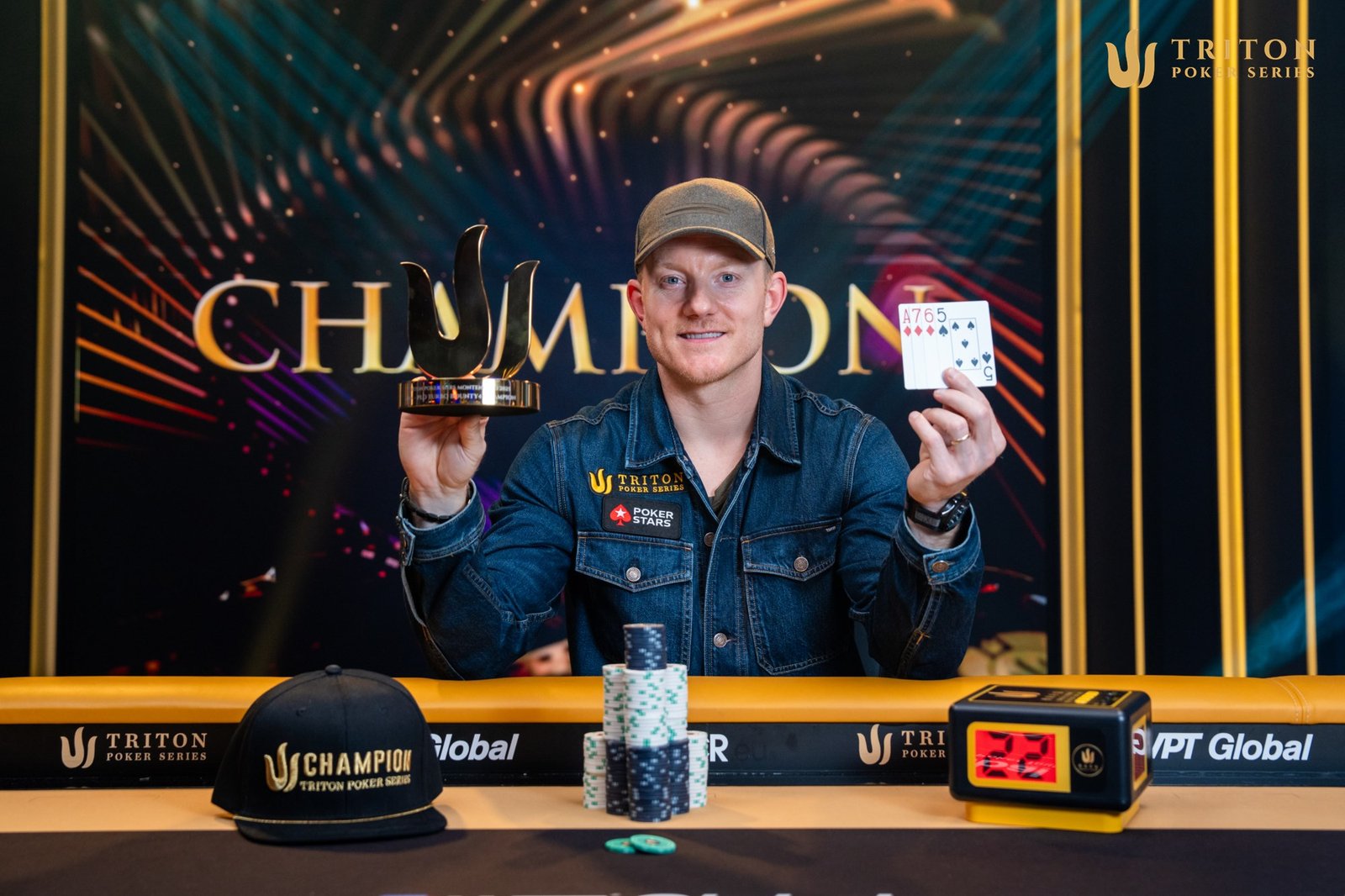 Ein Mann in einem dunklen Anzug und mit kurzem Haar sitzt an einem Pokertisch, umgeben von Spielkarten und Pokerchips. Er schaut konzentriert auf seine Karten und hat eine ernste Mi