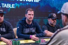Ein Mann mit dunklen Haaren und einem Bart sitzt an einem Pokertisch. Er trägt einen dunklen Anzug und hat eine ernste Miene.