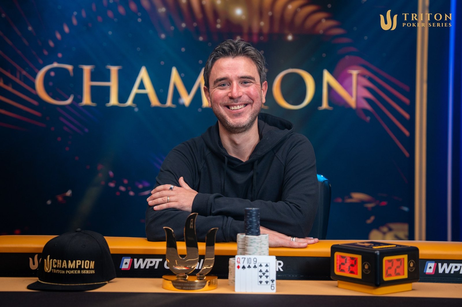 Ein Mann mit dunklem Haar und einem blauen Hemd sitzt lächelnd an einem Pokertisch in einem Casino. Er hält Karten in der Hand und schaut konzentriert auf den Tisch. Im H