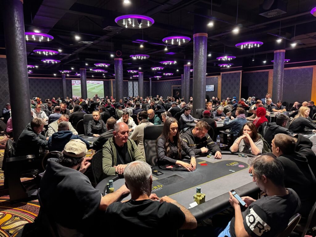 Ein voll besetzter Pokerraum in einem Casino, mit mehreren Tischen, an denen Spieler Karten spielen und Wetten platzieren. Die Atmosphäre ist lebhaft und spannend, mit vielen Menschen in Aktion