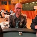 Ein professioneller Pokertisch mit grüner Filzoberfläche, umgeben von Stühlen mit Spielkarten und Pokerchips darauf. Im Hintergrund sind unscharfe Personen zu sehen, die sich auf das Spiel kon