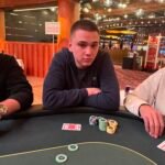 Ein professioneller Pokerspieler mit Sonnenbrille und Kappe sitzt an einem grünen Pokertisch und prüft seine Karten während eines Turniers.