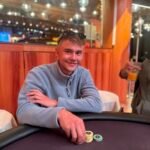 Ein Mann und eine Frau sitzen an einem Pokertisch und studieren konzentriert ihre Karten. Der Mann hat eine Hand am Kinn und die Frau hält einen Chip in der Hand. Im Hintergrund sind andere Spieler und