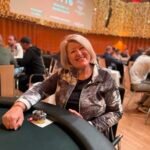 Ein grünes Pokertisch mit Spielkarten, Pokerchips und Getränken darauf, umgeben von Personen, die Karten halten und konzentriert spielen.