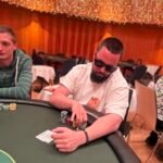 Ein Mann in einem schwarzen Anzug und einer Sonnenbrille spielt Poker an einem eleganten Holztisch in einem luxuriösen Casino, umgeben von anderen Spielern und einer glamourösen Atmosphäre.