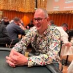 Ein spannendes Pokerspiel in einem eleganten Casino. Vier Personen sitzen um einen grünen Pokertisch, jeder hält Karten in der Hand und setzt Chips. Im Hintergrund sind weitere Spieler und eine