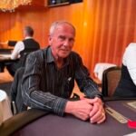 Ein Mann und eine Frau sitzen an einem Pokertisch, konzentriert auf ihre Karten und Chips. Die Frau hat blonde Haare und trägt eine rote Bluse, der Mann hat dunkle Haare und trägt ein