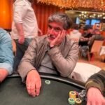 Ein Mann und eine Frau sitzen an einem Pokertisch und spielen Karten. Der Mann hat eine Pokerhand und schaut konzentriert auf seine Karten, während die Frau lächelnd ihre Karten betrachtet. Im