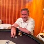 Ein Mann und eine Frau sitzen an einem Pokertisch und spielen eine Partie Poker. Der Mann hält Karten in der Hand, während die Frau konzentriert auf ihre eigenen Karten schaut. Chips und Getränke