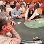 Ein Mann und eine Frau sitzen an einem Pokertisch und spielen eine Partie Poker. Der Mann hält Karten in der Hand und schaut konzentriert auf den Tisch, während die Frau mit gespanntem Blick ihre