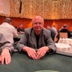 Ein Mann und eine Frau sitzen an einem Pokertisch und spielen gegeneinander. Der Mann hat eine Hand mit Karten in der Luft und schaut konzentriert auf die Spielkarten. Die Frau hat ebenfalls Karten in