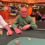 Ein Mann in einem Anzug und mit einer Sonnenbrille sitzt an einem Pokertisch und hält eine Hand mit Spielkarten. Ein Stapel Jetons liegt vor ihm.