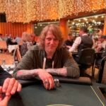 Ein Mann und eine Frau sitzen am Pokertisch und betrachten konzentriert ihre Karten. Die Frau hat blonde Haare und trägt ein schwarzes Kleid, der Mann hat dunkle Haare und trägt ein blaues