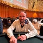 Ein Mann und eine Frau sitzen an einem Pokertisch und halten Karten in der Hand. Sie schauen konzentriert auf den Tisch. Im Hintergrund sind Casino-Chips und Getränke zu sehen.
