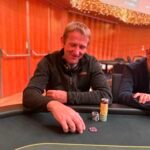 Ein Spieler mit Sonnenbrille und Kappe sitzt an einem Pokertisch und betrachtet seine Karten konzentriert, während er von anderen Spielern umgeben ist.
