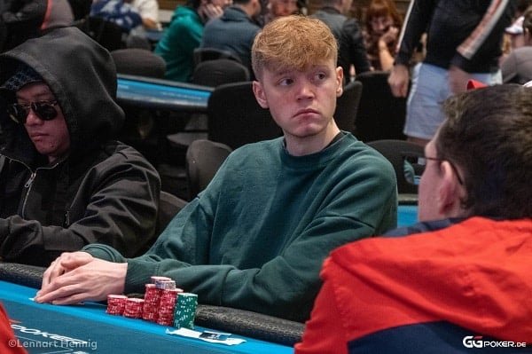 Ein Mann sitzt an einem Pokertisch und hält zwei Spielkarten in der Hand, während er konzentriert auf seine Gegner schaut.