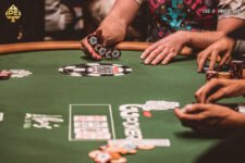 Ein Mann sitzt an einem Pokertisch mit einem großen Stapel Pokerchips vor sich. Er hat eine Sonnenbrille auf und betrachtet seine Karten mit einem konzentrierten Blick.