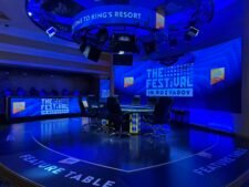 Ein Mann sitzt an einem Pokertisch mit Pokerchips und Karten vor sich. Er hat eine nachdenkliche Miene und scheint konzentriert auf sein Blatt zu schauen. Im Hintergrund sind weitere Spieler