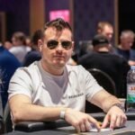 Ein Mann mit schwarzer Sonnenbrille und einem roten Hemd hält zwei Spielkarten in der Hand und schaut konzentriert auf den Pokertisch. Im Hintergrund sind Chips und weitere Spieler zu sehen.