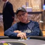 Ein Mann mit dunklen Haaren und einem blauen Hemd sitzt an einem Pokertisch und hält zwei Spielkarten in der Hand. Er schaut konzentriert auf den Tisch, umgeben von Chips und anderen Spiel