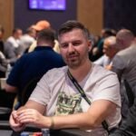 Ein Mann und eine Frau sitzen an einem Pokertisch und spielen. Der Mann hält Karten in der Hand, während die Frau in Gedanken versunken scheint. Im Hintergrund sind Pokerchips und Getränke zu