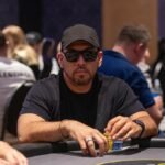 Ein Mann in einem dunkelblauen Anzug sitzt an einem Pokertisch und hält zwei Karten in der Hand. Er trägt eine Sonnenbrille und starrt konzentriert auf seine Karten. Im H